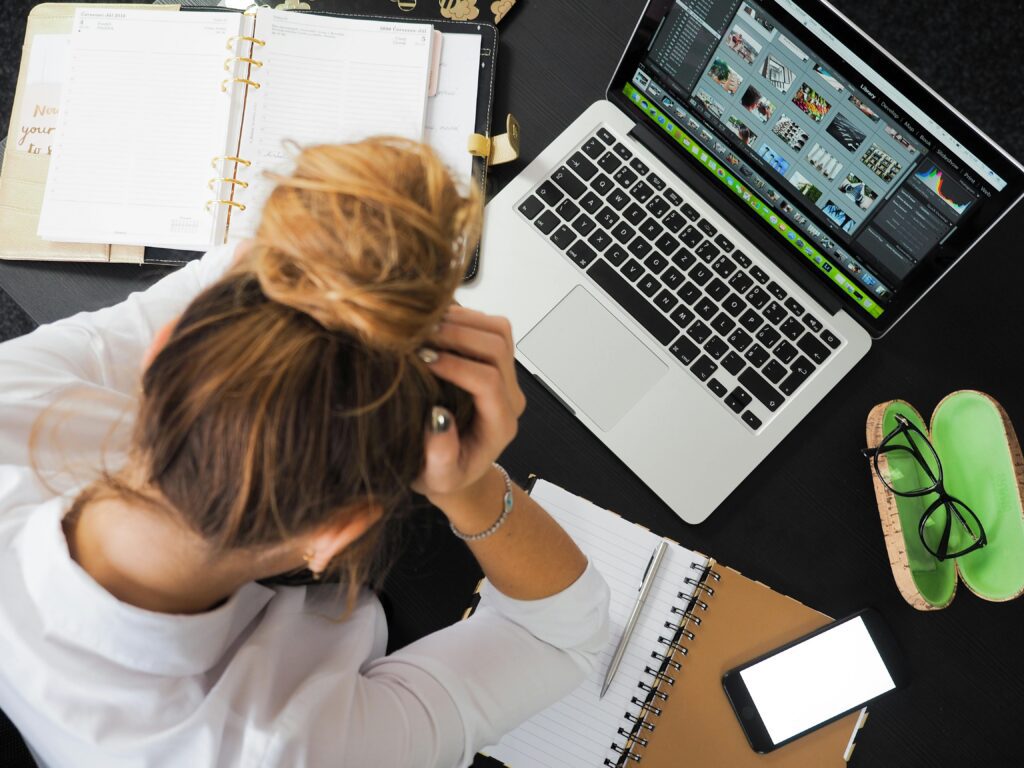 a lady with her head over her desk looking overwhelmed by paperwork and a laptop screen. 